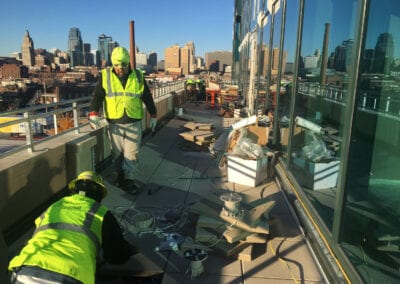 group of construction workers is engaged in a project on the rooftop of a building in Kansas City. The workers are wearing high-visibility vests, hard hats, and gloves for safety. They are handling various construction materials and tools spread out on the rooftop. One worker is kneeling, possibly installing or adjusting tiles or other elements of the rooftop. The background showcases the Kansas City skyline, with its distinctive buildings and clear blue sky. The scene highlights the ongoing construction work and the urban setting, with reflections of the cityscape visible on the building's glass facade.