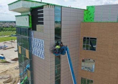 a team of workers is performing architectural sheet metal work on the side of a tall building in Kansas City. The workers are on a boom lift, which allows them to reach high sections of the building's exterior safely. They are equipped with safety gear, including hard hats and harnesses, and are handling metal panels or sheets, likely installing or repairing them. The background shows the impressive height of the building and the surrounding urban landscape, emphasizing the scale and complexity of the task