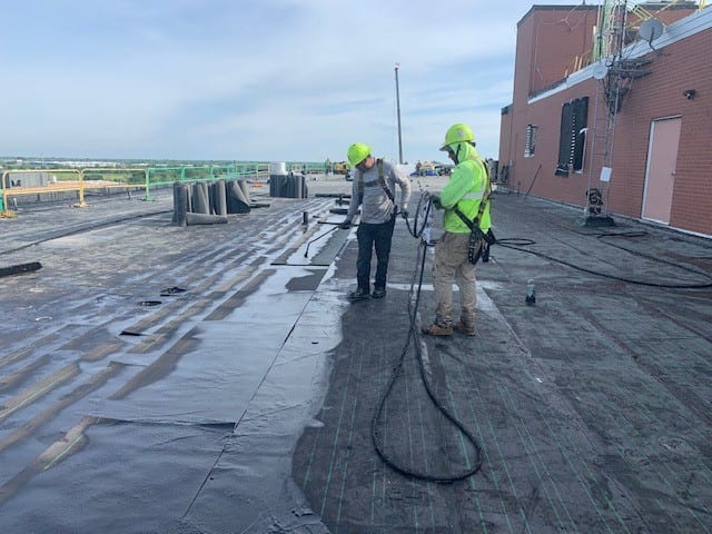 several workers are performing maintenance on a commercial roof in Kansas City. They are equipped with safety gear, including hard hats and high-visibility vests. The workers are spread out across the roof, using various tools and materials to ensure the roof is in good condition. The background shows a clear sky and the surrounding urban landscape, highlighting the location and the extent of the maintenance work