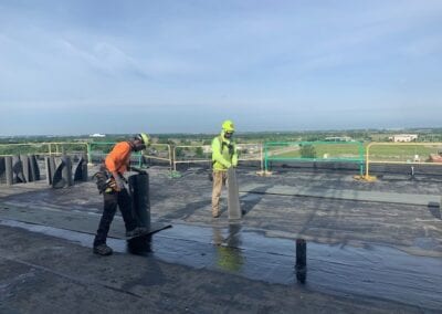 several workers are seen performing maintenance on a commercial roof. They are equipped with safety gear, including harnesses, hard hats, and high-visibility vests. The workers appear to be inspecting and repairing various sections of the roof, ensuring it is in good condition. The roof has multiple tools and materials spread out, indicating ongoing work. The background shows part of the building and an expansive view of the surrounding Kansas City area, highlighting the elevated work environment.