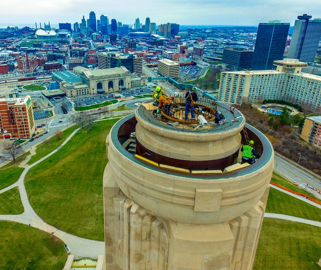 Delta Services commercial roofing company employees performing maintenance or construction work on the top of the National WWI Museum and Memorial in Kansas City. The workers are wearing safety gear, including hard hats, harnesses, and high-visibility vests, and they are using various tools and equipment. They are positioned around the circular top section of the structure, which appears to be a lookout or observation platform. The background showcases the Kansas City skyline, with notable buildings and landmarks visible, as well as the surrounding park and urban landscape. The height and perspective emphasize the impressive elevation and the complexity of the task the workers are undertaking.