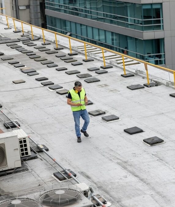 a worker is conducting a commercial roof inspection on the rooftop of a building. The worker is wearing a high-visibility vest and safety gear while carrying a clipboard or tablet to document findings. The rooftop features various HVAC units and other equipment, with safety railings along the edges. The surrounding buildings are modern and tall, indicating an urban setting. The worker is meticulously walking across the roof, inspecting for any issues or necessary repairs