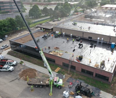 a large commercial roof repair project is underway on a flat-roofed building. Numerous workers are spread out across the roof, engaged in various tasks such as removing old roofing material, preparing the surface, and installing new materials. A crane is positioned next to the building, lifting materials and equipment onto the roof. Several vehicles and pieces of heavy machinery are also visible around the site, indicating a well-coordinated operation. The surrounding area includes other buildings, trees, and streets, providing context to the urban setting of the project