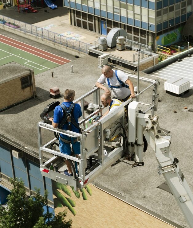 a team of workers is performing a commercial roof inspection using a boom lift. The workers are wearing safety harnesses and high-visibility vests as they inspect the rooftop of a large building. The building appears to be part of a school or an institution, as indicated by the adjacent playground and sports court. The workers are closely examining the roof, possibly looking for signs of damage or wear. The surrounding area includes multiple buildings, indicating an urban environment